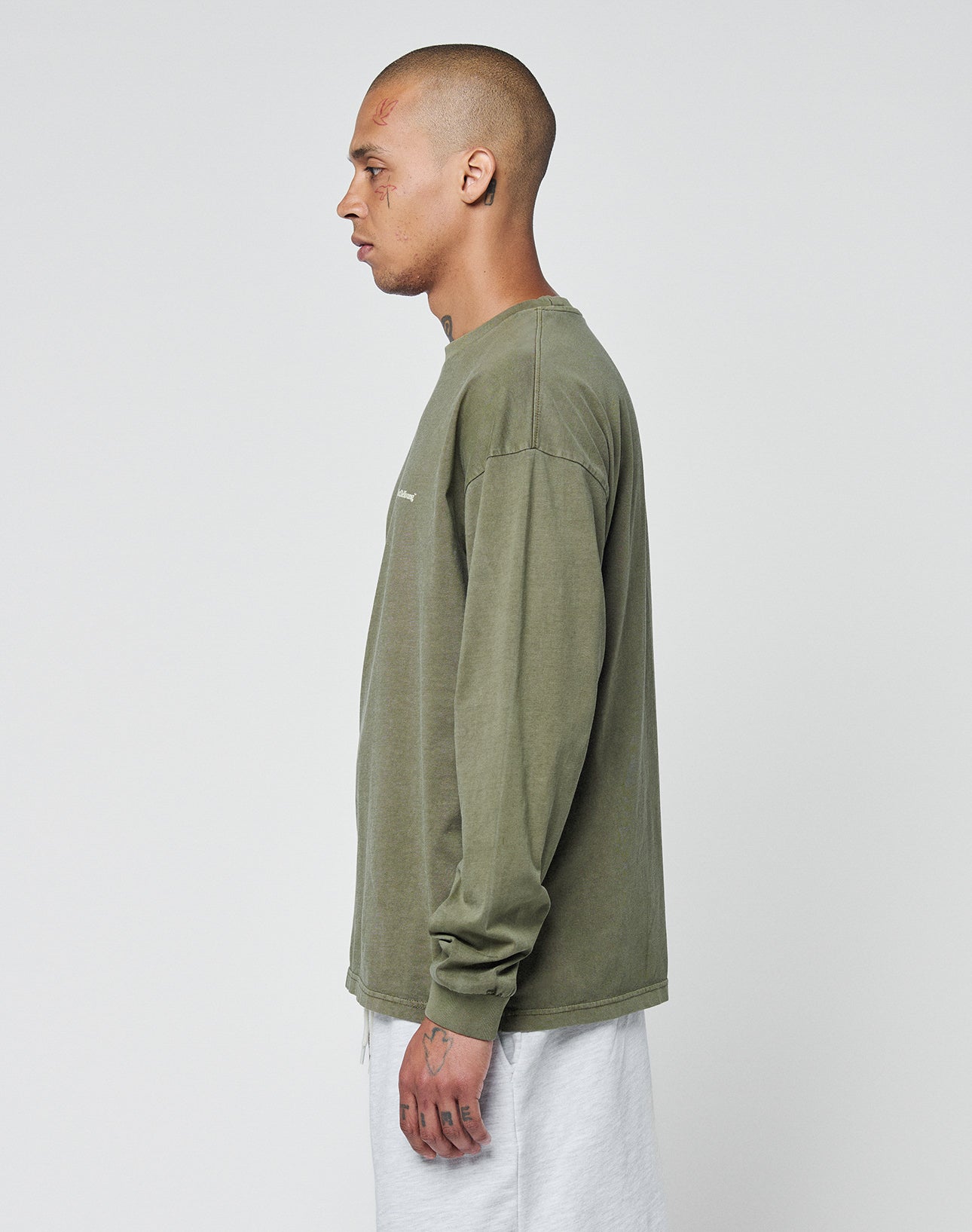 A person with a shaved head and face tattoos stands in profile wearing the LIVE FAST DIE YOUNG Basic Longsleeve in oversize fit, olive green 100% Baumwolle, paired with light gray pants against a white background.