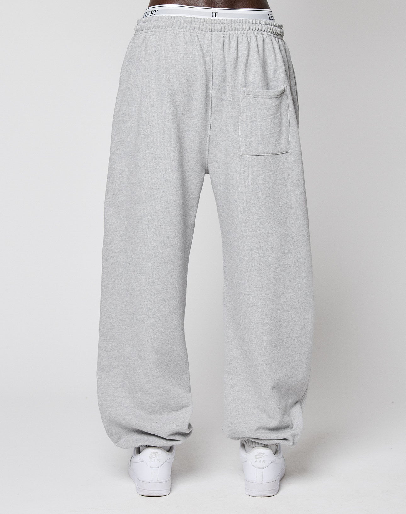 A person wears the LIVE FAST DIE YOUNG Wide Cuff Jogger in light gray French Terry with an Elastikbund waistband and front pocket, styled with white sneakers, standing against a plain white background.
