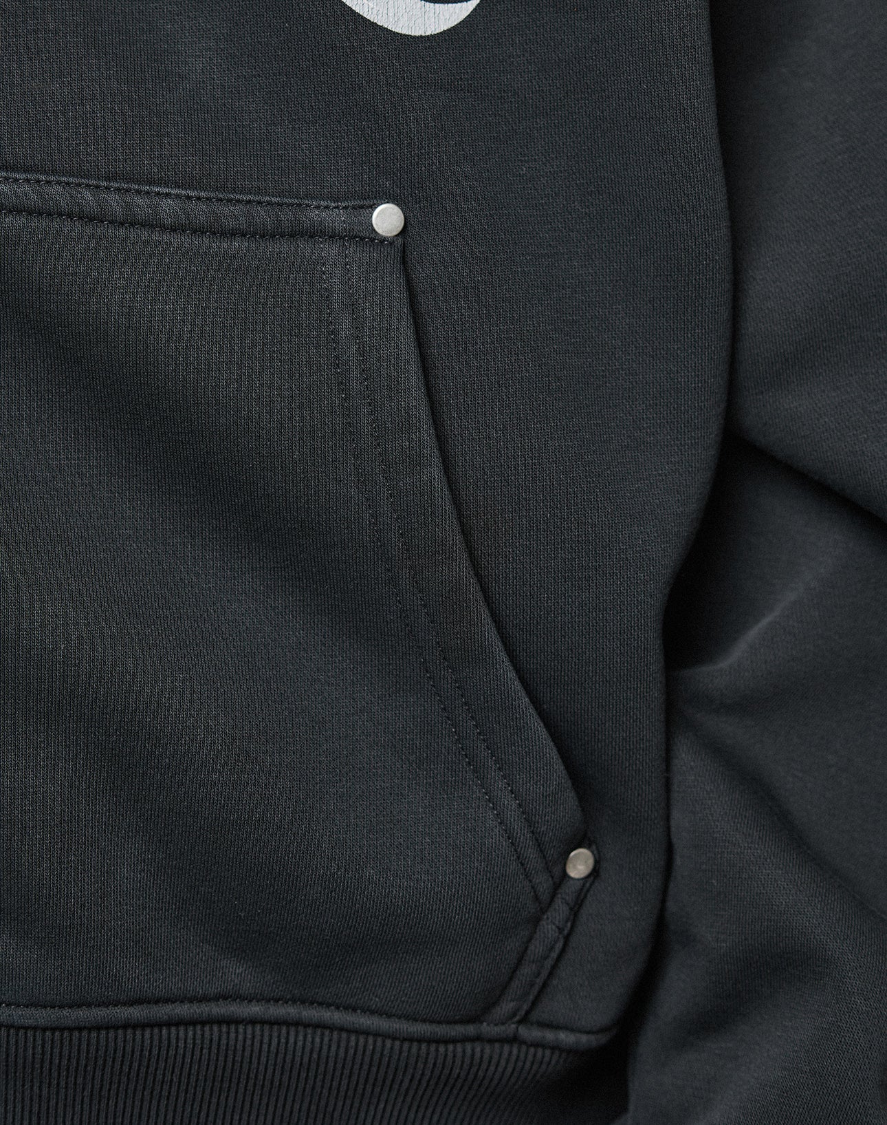 Close-up of the black LFDY Inferno Hooded Zip by LIVE FAST DIE YOUNG, showing its oversize pocket with visible stitching and three metallic rivets. The edge of a white front print hints at the individueller Wascheffekt on the fabric.