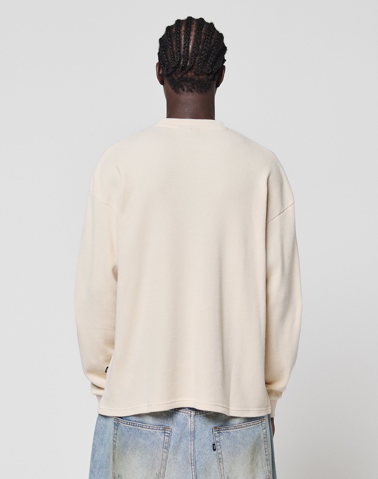 Seen from behind, a person with cornrow braids wears the LIVE FAST DIE YOUNG Blank Waffle Longsleeve—an oversize, cream 100% Baumwolle shirt—paired with light blue jeans, standing against a plain light background.