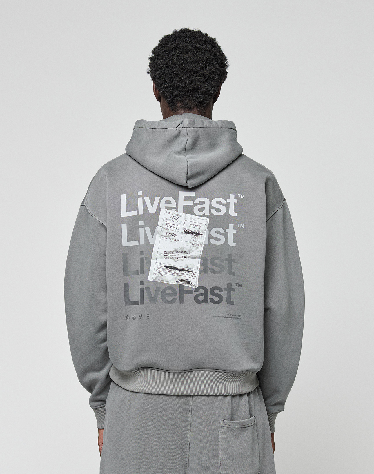 A person wears the Basic Evidence Hooded by LIVE FAST DIE YOUNG, an oversize gray hoodie with “Live Fast” repeated in white and a crumpled paper graphic on the back, shown from behind. | model