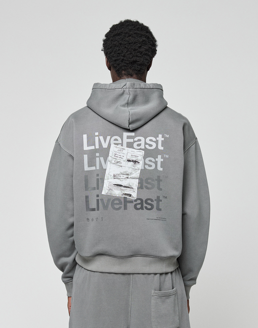 A person wears the Basic Evidence Hooded by LIVE FAST DIE YOUNG, an oversize gray hoodie with “Live Fast” repeated in white and a crumpled paper graphic on the back, shown from behind. | model