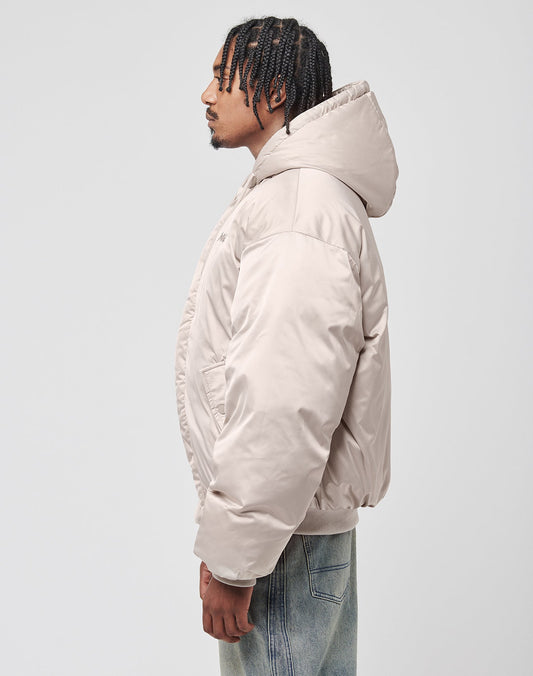 A man with braided hair, in profile, wears the LIVE FAST DIE YOUNG LFDY Winter Bomber Jacket in beige with hood and oversize fit, paired with light blue jeans, standing against a plain light gray background.