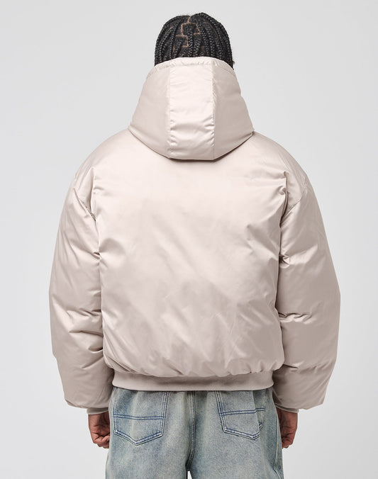 A person with braided hair is facing away, wearing the LFDY Winter Bomber Jacket by LIVE FAST DIE YOUNG in light beige with oversize fit and hood, paired with light blue jeans against a plain light background.