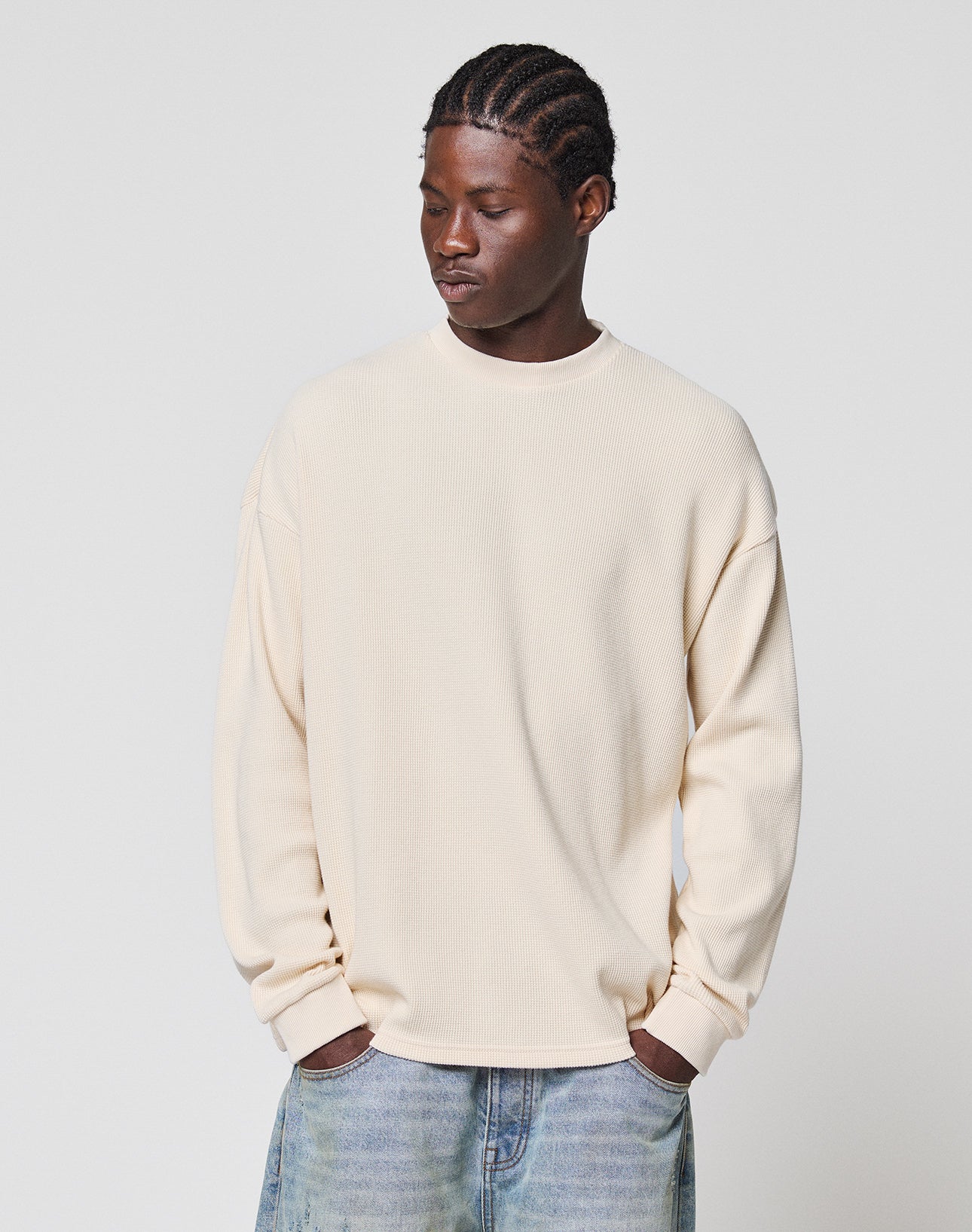 A man with braided hair looks down with his hands in his pockets, wearing the LIVE FAST DIE YOUNG Blank Waffle Longsleeve in an oversize fit and light blue 100% Baumwolle jeans, standing against a plain light gray background.