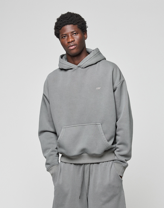 A young man with short curly hair wears the LIVE FAST DIE YOUNG Basic Evidence Hooded gray hoodie and matching pants, standing against a plain light background with his hands in his pockets, looking at the camera with a slight smile.