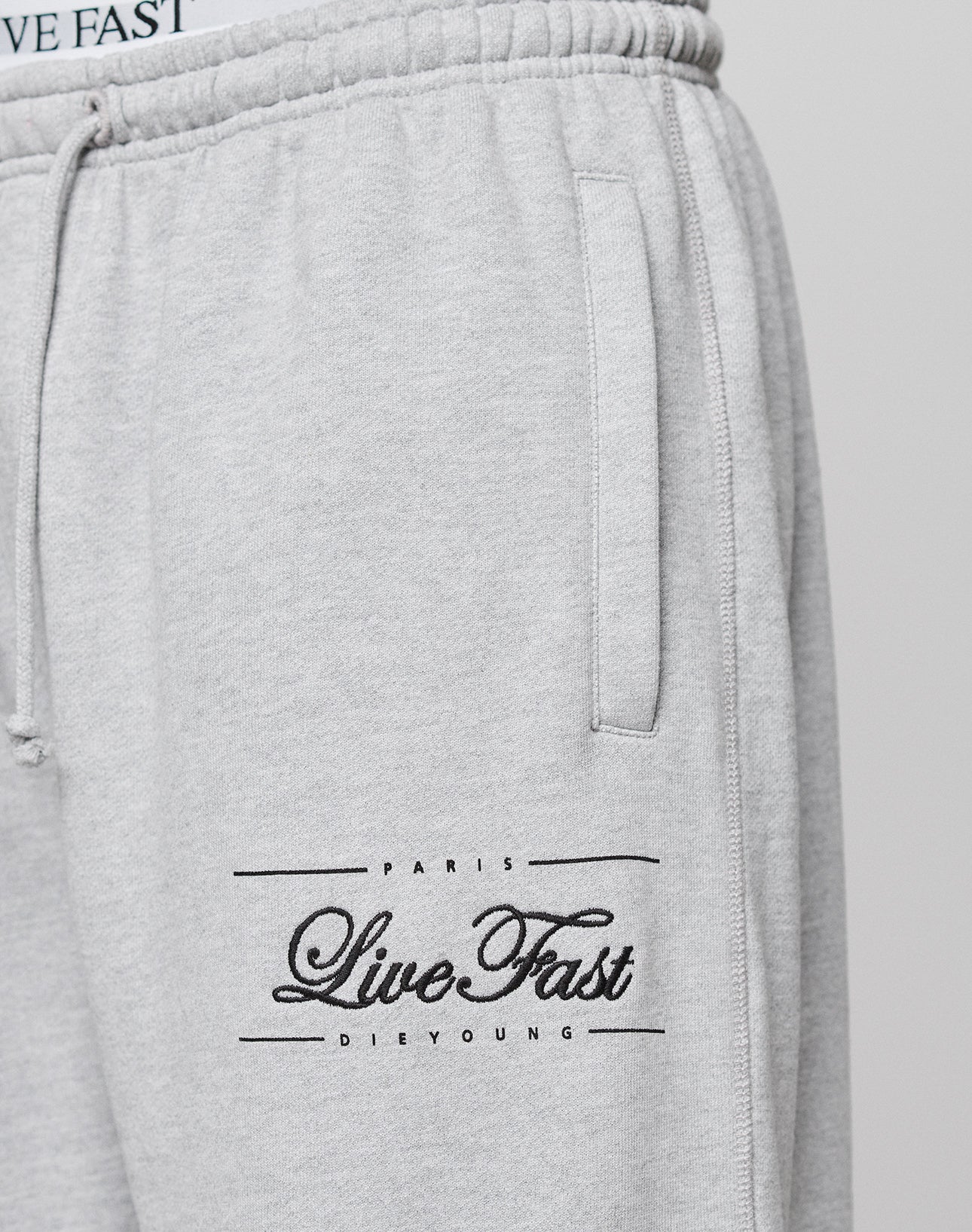 Close-up of the Wide Cuff Jogger by LIVE FAST DIE YOUNG in light grey French Terry, featuring a wide fit, elastic waistband, side pocket, and "PARIS LIVE FAST DIE YOUNG" text in black on the left thigh.
