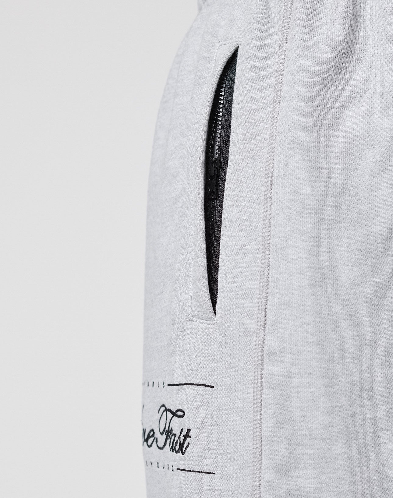 Close-up of the LIVE FAST DIE YOUNG Wide Cuff Jogger, a wide fit, light grey French Terry sweatpant with a black zippered pocket and partial black text near the pocket, shown on a plain background.