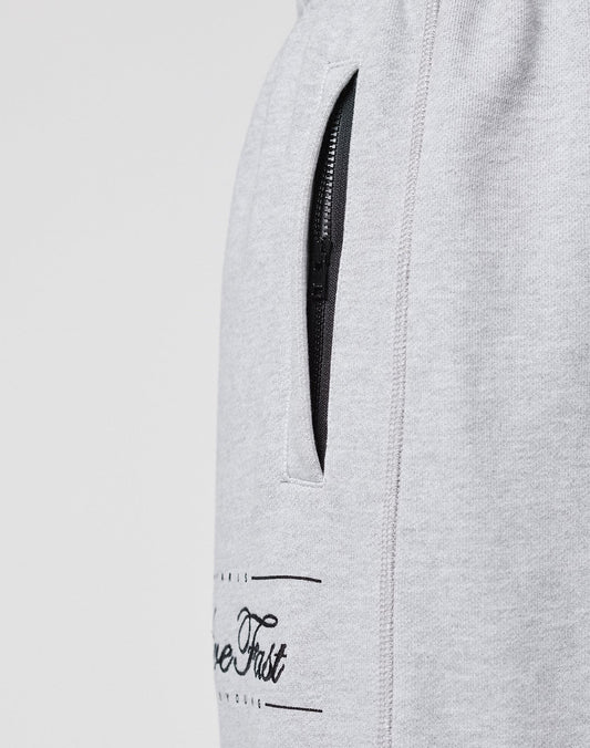 Close-up of the LIVE FAST DIE YOUNG Wide Cuff Jogger, a wide fit, light grey French Terry sweatpant with a black zippered pocket and partial black text near the pocket, shown on a plain background.