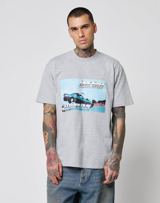 A tattooed man wears the LFDY Crash Kid Tee by LIVE FAST DIE YOUNG—a regular fit, light gray graphic T-shirt with a car and Live Fast text—standing against a plain, light background.