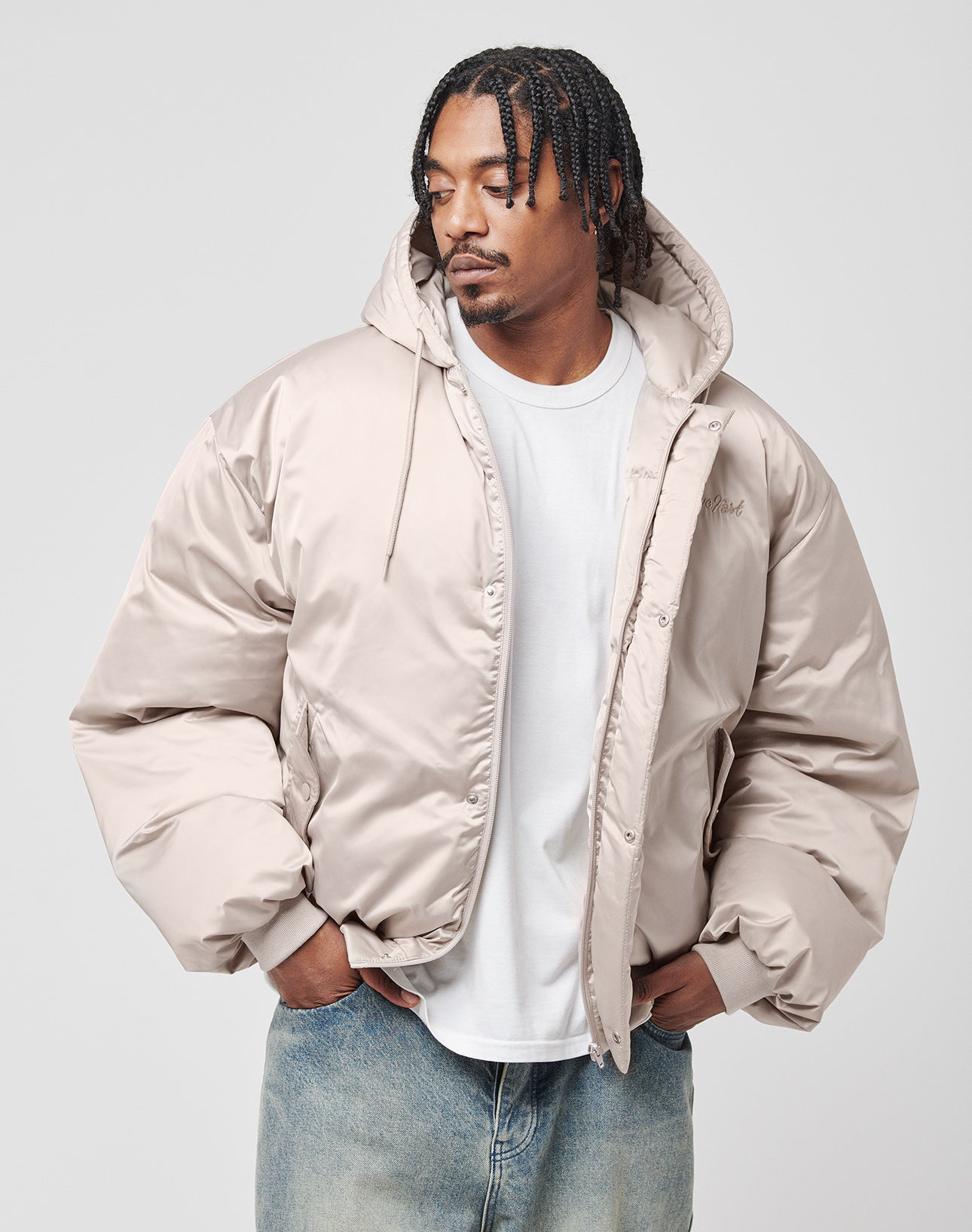 A man with braided hair wears the LIVE FAST DIE YOUNG LFDY Winter Bomber Jacket in light beige, styled oversize over a white T-shirt and blue jeans, standing with hands in pockets and looking left against a plain light background.
