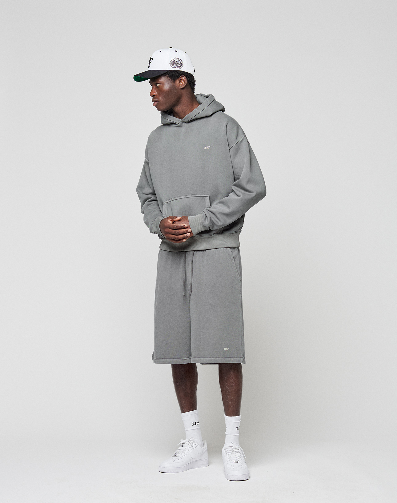 A person wears LIVE FAST DIE YOUNG Basic Shorts and a gray hoodie with a subtle logo, along with white socks, sneakers, and a white baseball cap, standing against a plain light gray background.