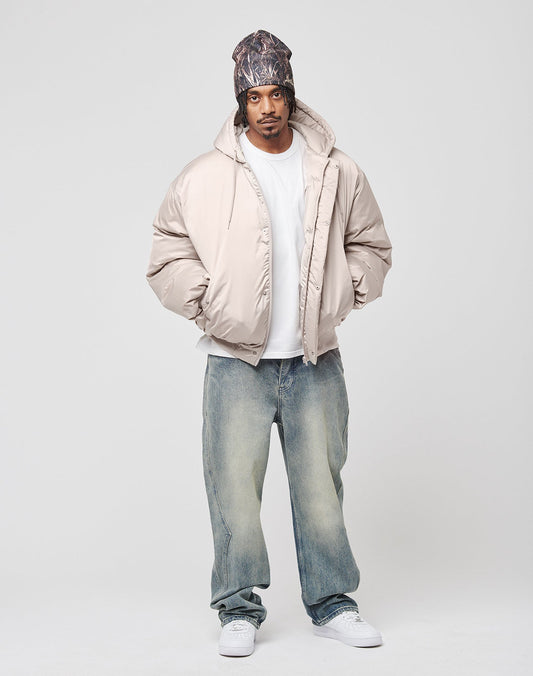 A man in a beige LFDY Winter Bomber Jacket by LIVE FAST DIE YOUNG, white t-shirt, faded baggy jeans, white sneakers, and a patterned beanie stands facing forward against a plain light background.