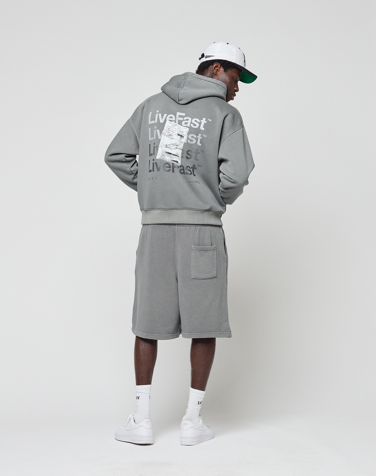 A person in an oversize fit gray Basic Evidence Hooded by LIVE FAST DIE YOUNG, featuring a faded finish and repeated "Live Fast" text on the back, stands facing away. They wear shorts, white sneakers, socks, and a cap against a plain white background.