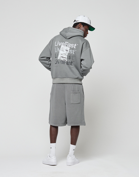 A person in an oversize fit gray Basic Evidence Hooded by LIVE FAST DIE YOUNG, featuring a faded finish and repeated "Live Fast" text on the back, stands facing away. They wear shorts, white sneakers, socks, and a cap against a plain white background.