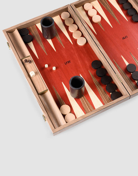 The LFDY Backgammon by LIVE FAST DIE YOUNG is a limited edition wooden board handcrafted in Greece, featuring walnut playing pieces, dice, and cups on a red and tan surface with light and dark pieces set up on opposite sides.