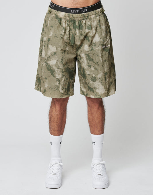 A person wears LIVE FAST DIE YOUNGs Live Fast Nylon Shorts in green camo with side pockets, paired with LIV text white socks, white sneakers, and black waistband underwear reading LIVE FAST. Photo is waist-down against a plain backdrop.
