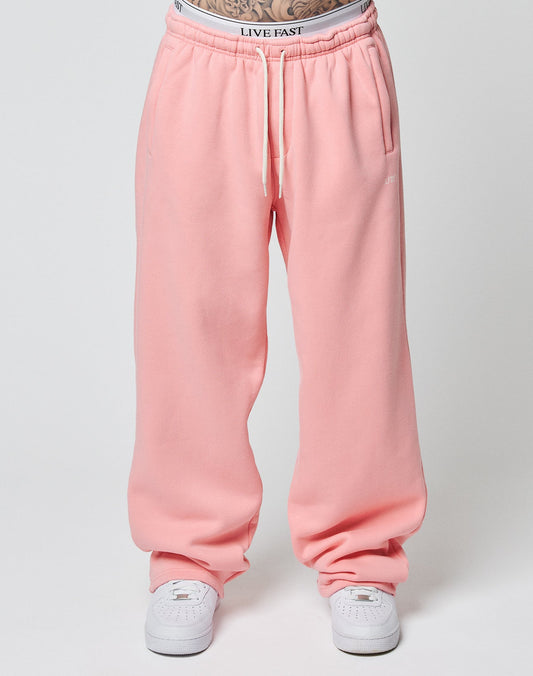 Person wears loose-fitting pink Basic Wide Jogger sweatpants by LIVE FAST DIE YOUNG with an elastic drawstring waist and a white logo waistband, paired with white sneakers. Image is cropped above the waist; plain, light background.
