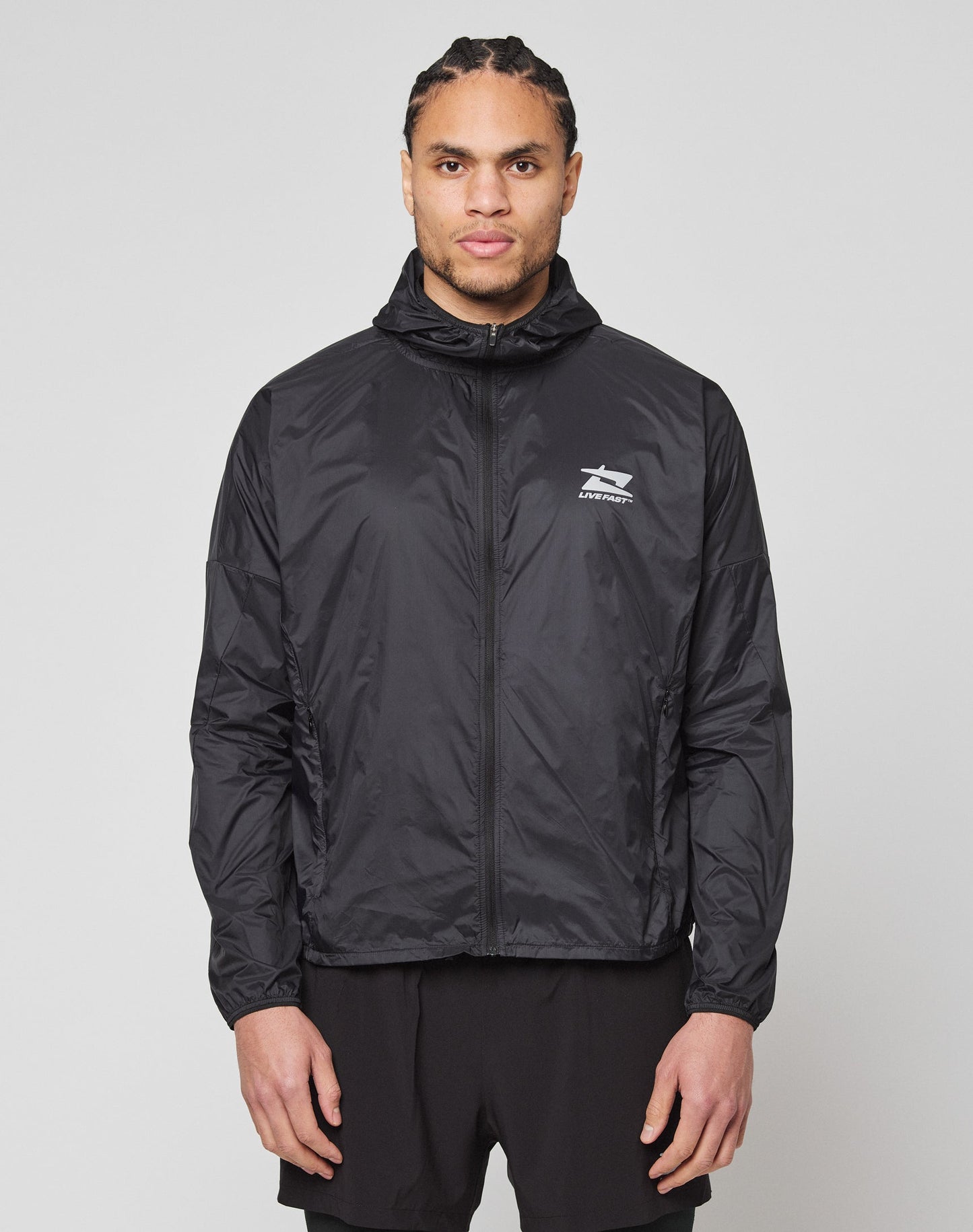 A man with neatly braided hair stands facing forward in a LIVE FAST DIE YOUNG Live Fast Performance Running Jacket, featuring a black zip-up design with Kapuze mit Visor and reflektierender Frontprint, plus black shorts, against a plain light gray background. | model