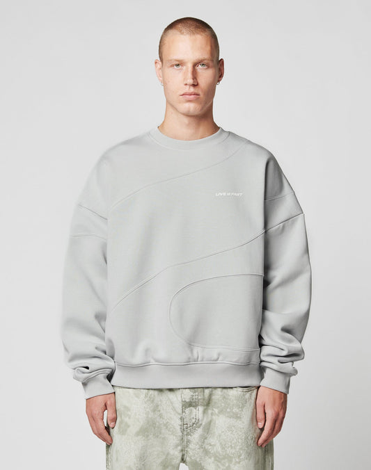 A person with short hair wears the LIVE FAST DIE YOUNG LFDY Wavy Sweater in light gray Oversize Fit, showcasing curved seam details and small white chest text, paired with light gray-green pants against a plain light background.