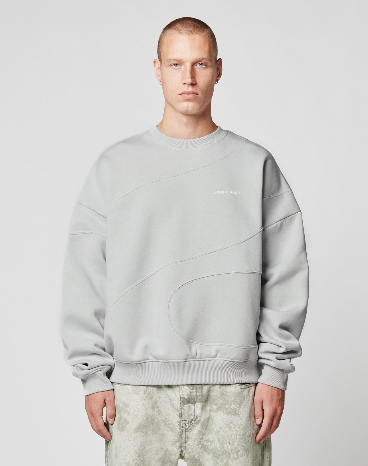 A person with short hair wears the LIVE FAST DIE YOUNG LFDY Wavy Sweater in light gray Oversize Fit, showcasing curved seam details and small white chest text, paired with light gray-green pants against a plain light background.