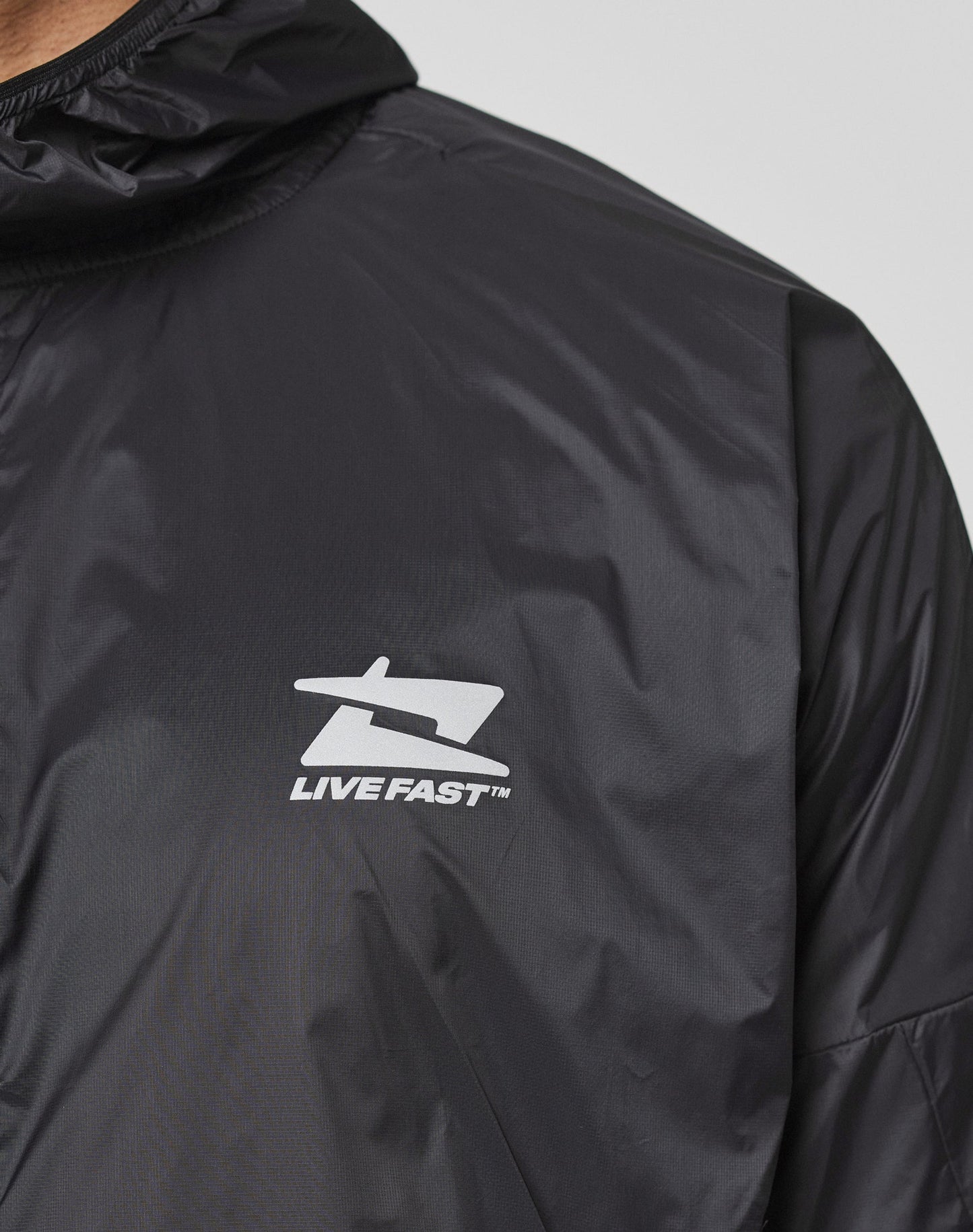 Close-up of the Live Fast Performance Running Jacket by LIVE FAST DIE YOUNG, featuring a black design with a white LIVE FAST logo and stylized chest graphic. Shiny texture, Kapuze mit Visor, Regular Fit, and reflektierender Frontprint for standout style.