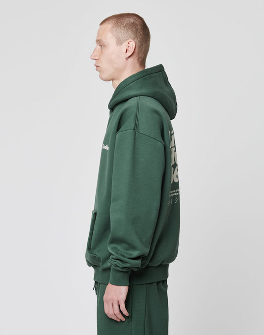A person with short hair stands in profile against a light gray background, wearing the LIVE FAST DIE YOUNG Basic 360 Hooded in dark green with an oversized fit, front and back print, matching pants, and a front pocket.