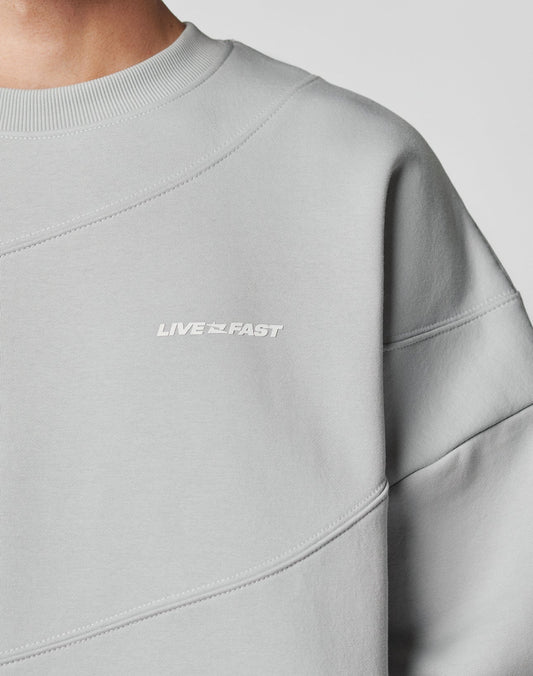 Close-up of the LFDY Wavy Sweater by LIVE FAST DIE YOUNG—a light gray, oversize-fit sweatshirt made from soft cotton with LIVE FAST in small white text on the chest. Only part of the wearers neck and shoulder are shown.