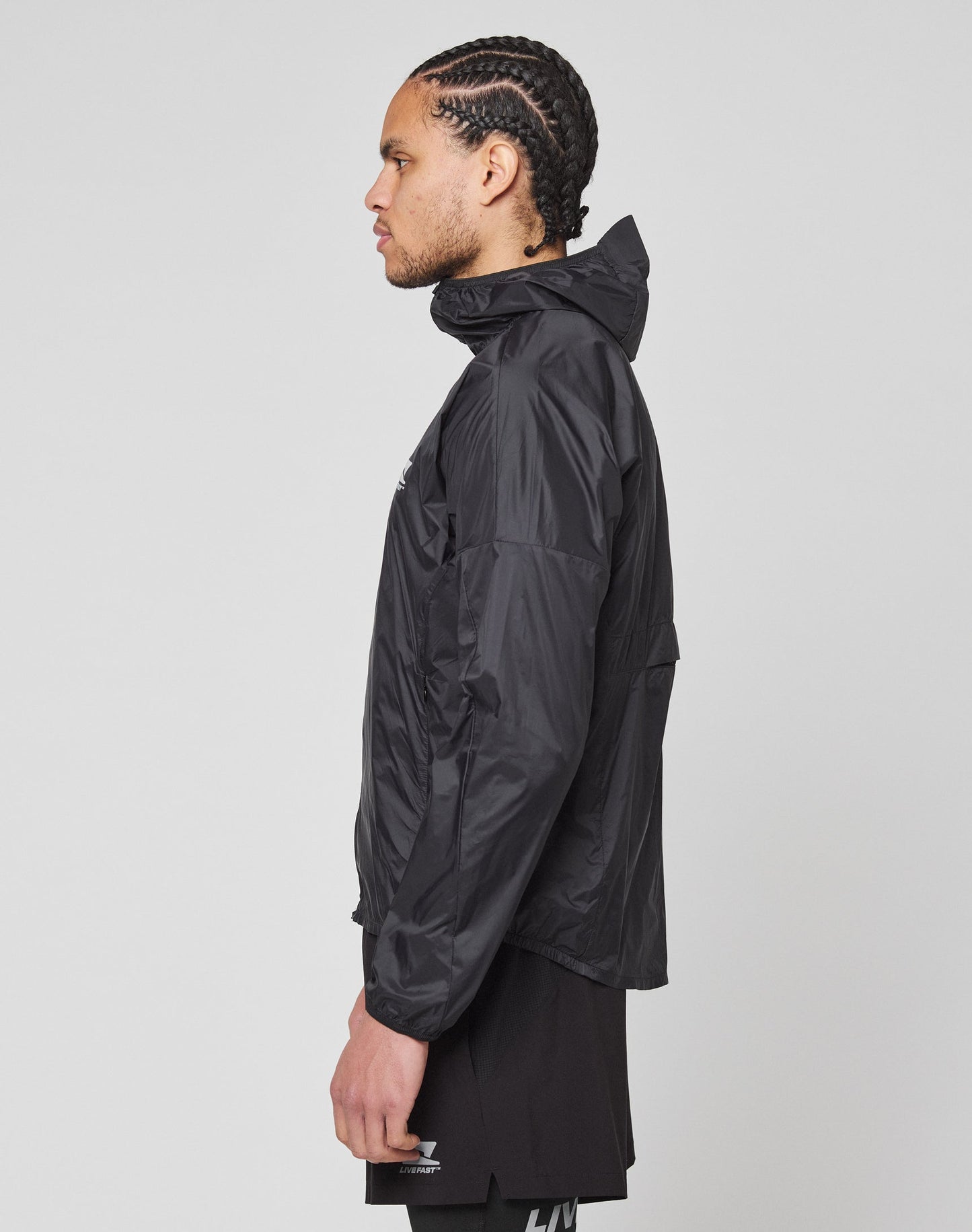 A person with braided hair stands in profile against a plain light gray background, wearing the LIVE FAST DIE YOUNG Live Fast Performance Running Jacket in black and black shorts, featuring a reflective front print.