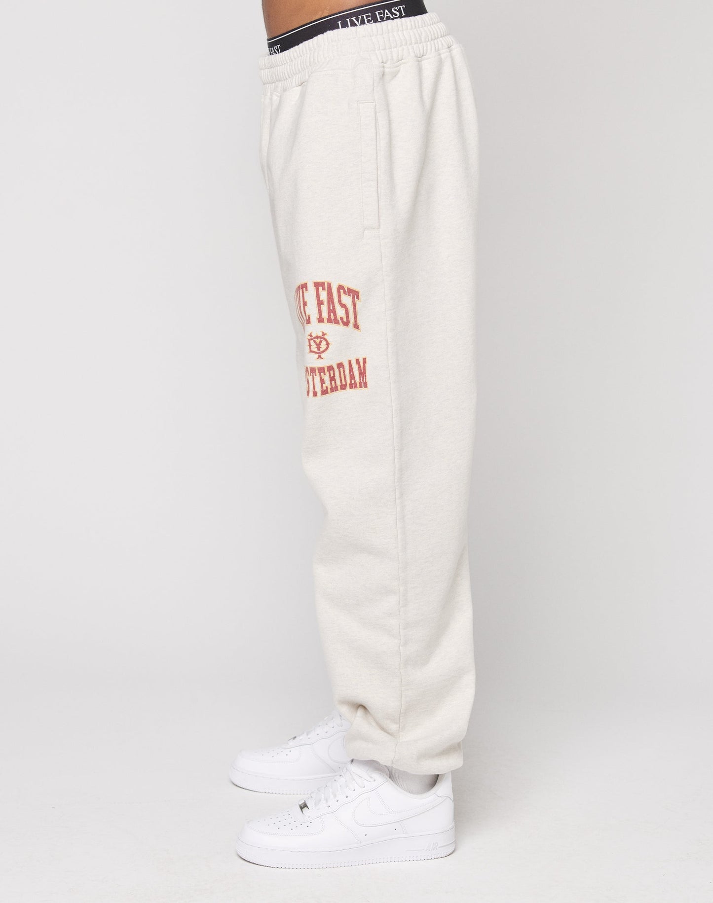 Person trägt Live Fast Amsterdam Loose Jogger von LIVE FAST DIE YOUNG in Off-White mit Elastikbund und rotem Schenkel-Schriftzug, kombiniert mit weißen Sneakers im Oversize Fit vor weißem Hintergrund.