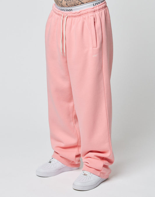 A person wearing LIVE FAST DIE YOUNG Basic Wide Jogger sweatpants in pink with white drawstrings and white sneakers, standing against a plain light background. Only their lower body is visible. | model