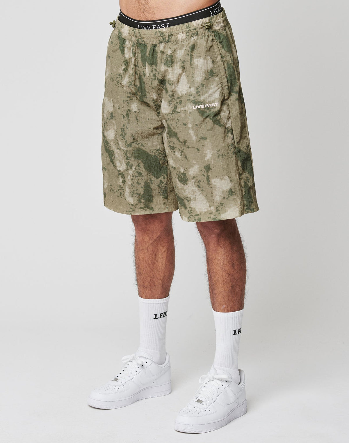 Someone stands against a plain light background wearing LIVE FAST DIE YOUNG Live Fast Nylon Shorts in green and beige camo with lateral pockets and silicone LIVE FAST prints, paired with white crew socks and white sneakers. | model