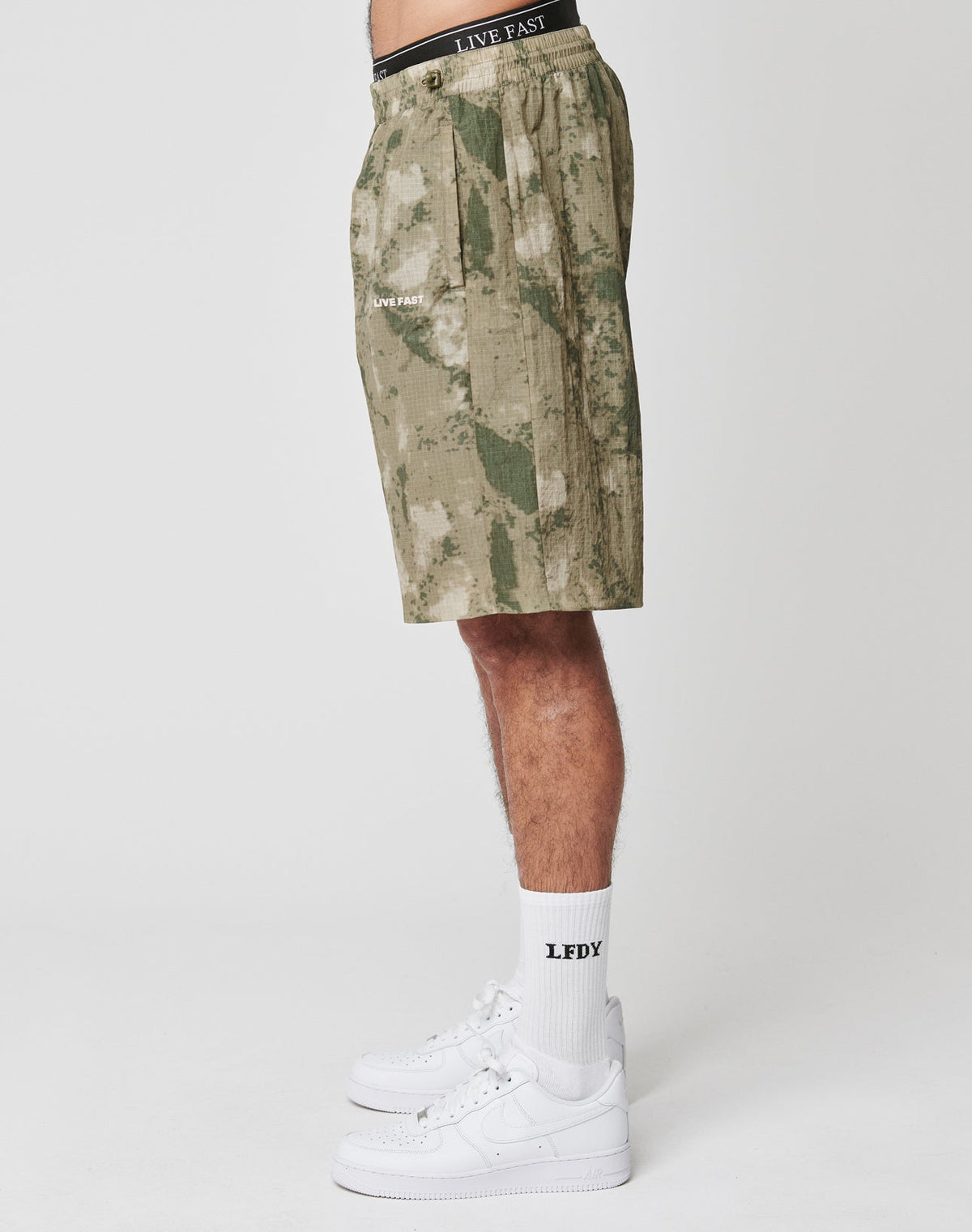 A person in LIVE FAST DIE YOUNG Live Fast Nylon Shorts with side pockets and LIVE FAST silicone print, paired with white LFDX socks and white sneakers, shown in side profile against a plain white background.