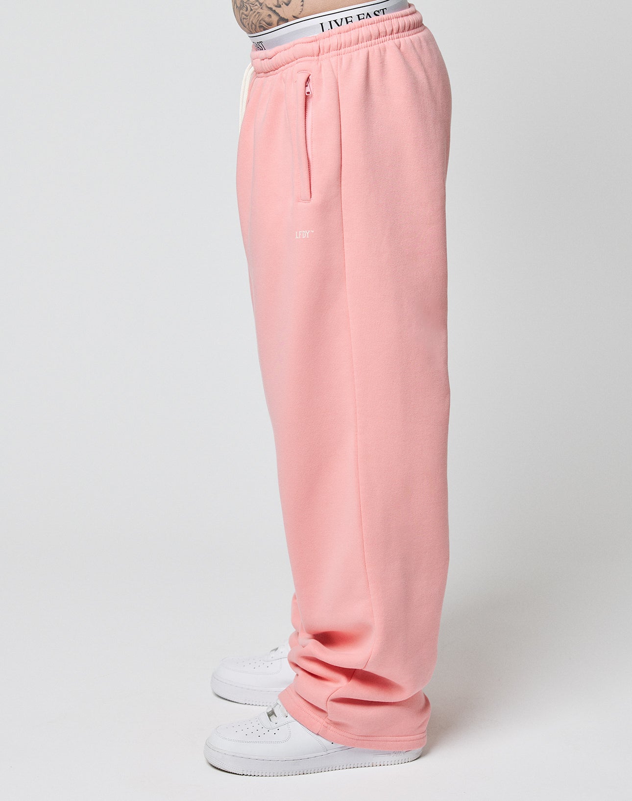 A person wearing LIVE FAST DIE YOUNGs Basic Wide Jogger in light pink with an elastic waistband and white sneakers, standing sideways against a plain white background.
