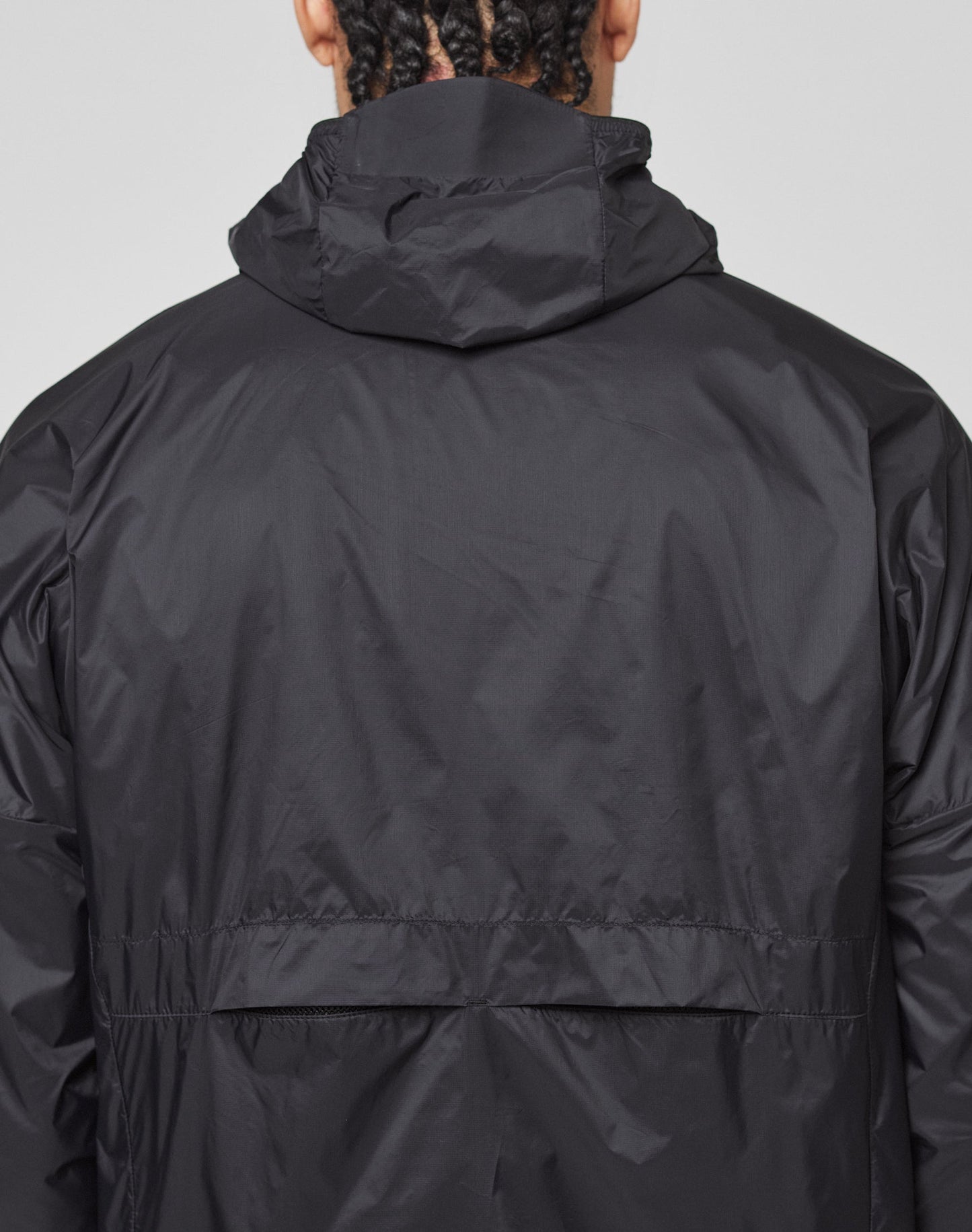 A person with braided hair, seen from behind, wears the LIVE FAST DIE YOUNG Live Fast Performance Running Jacket in black with a visor hood, shiny lightweight fabric, and a reflective front print.