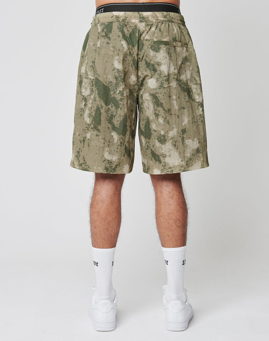 A person faces away from the camera wearing LIVE FAST DIE YOUNG’s Live Fast Nylon Shorts in green camo, paired with white socks and sneakers. The plain background is white.