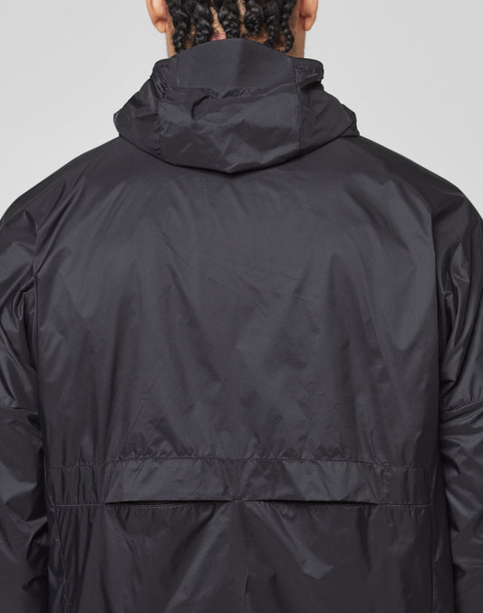 A person with braided hair, seen from behind, wears the LIVE FAST DIE YOUNG Live Fast Performance Running Jacket in black with a visor hood, shiny lightweight fabric, and a reflective front print.