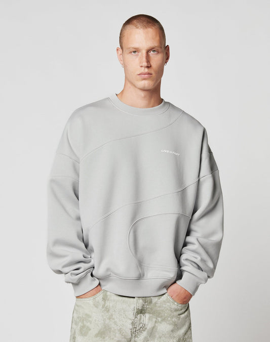 A young man with short hair wears the LIVE FAST DIE YOUNG LFDY Wavy Sweater, a loose light gray Baumwolle sweatshirt with curved stitching and chest text, paired with oversize light gray pants, standing against a plain white background.