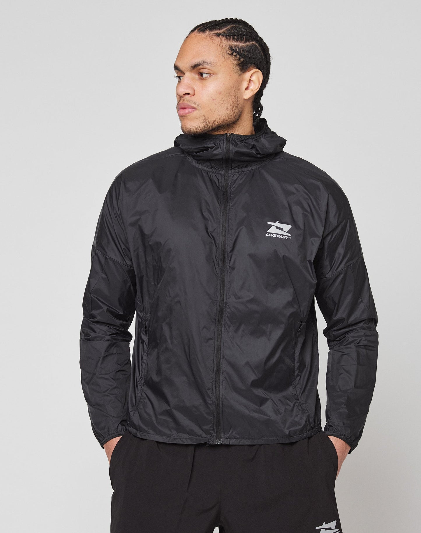 A man in a black Live Fast Performance Running Jacket by LIVE FAST DIE YOUNG, featuring a hood with visor and reflective front print, stands against a plain light background, looking left with his hands in his pockets.