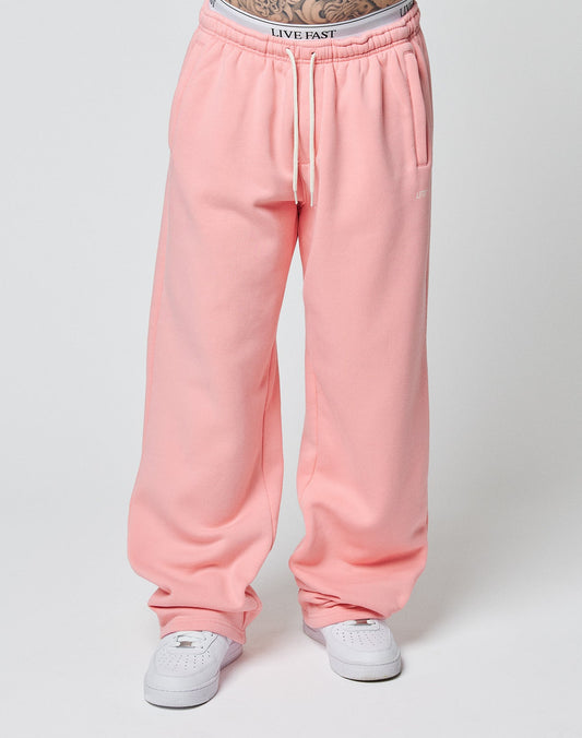 A person wears the LIVE FAST DIE YOUNG Basic Wide Jogger in loose pink with an elastic waistband, side pockets, and white drawstring. LIVE FAST underwear is visible above the pants. The white sneakers are set against a plain light background.