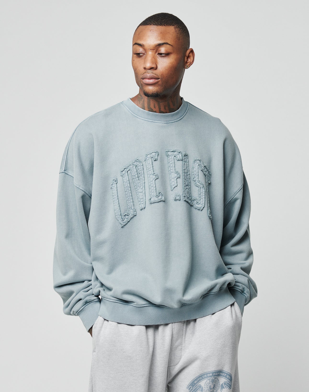 A man stands against a plain light background, looking down with his hands in his pockets, wearing the LIVE FAST DIE YOUNG Ripped College Sweater in light blue with distressed lettering and paired with light gray sweatpants.