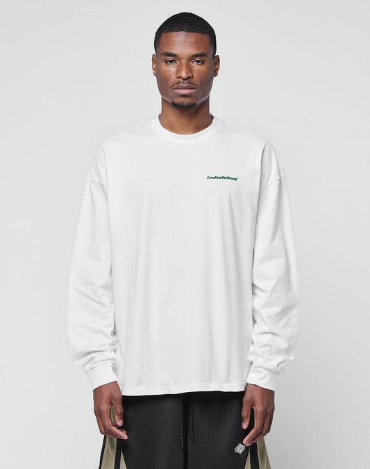 A man faces forward in a LIVE FAST DIE YOUNG Basic Longsleeve, an oversized white cotton shirt with a small green logo print on the chest, paired with black shorts featuring tan side panels, against a plain light gray background.| model