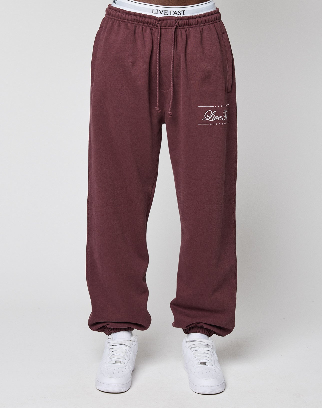 Someone wearing maroon Wide Cuff Jogger sweatpants by LIVE FAST DIE YOUNG, featuring "Live Fast" on the left thigh and branded waistband, paired with white sneakers, stands against a plain light background.