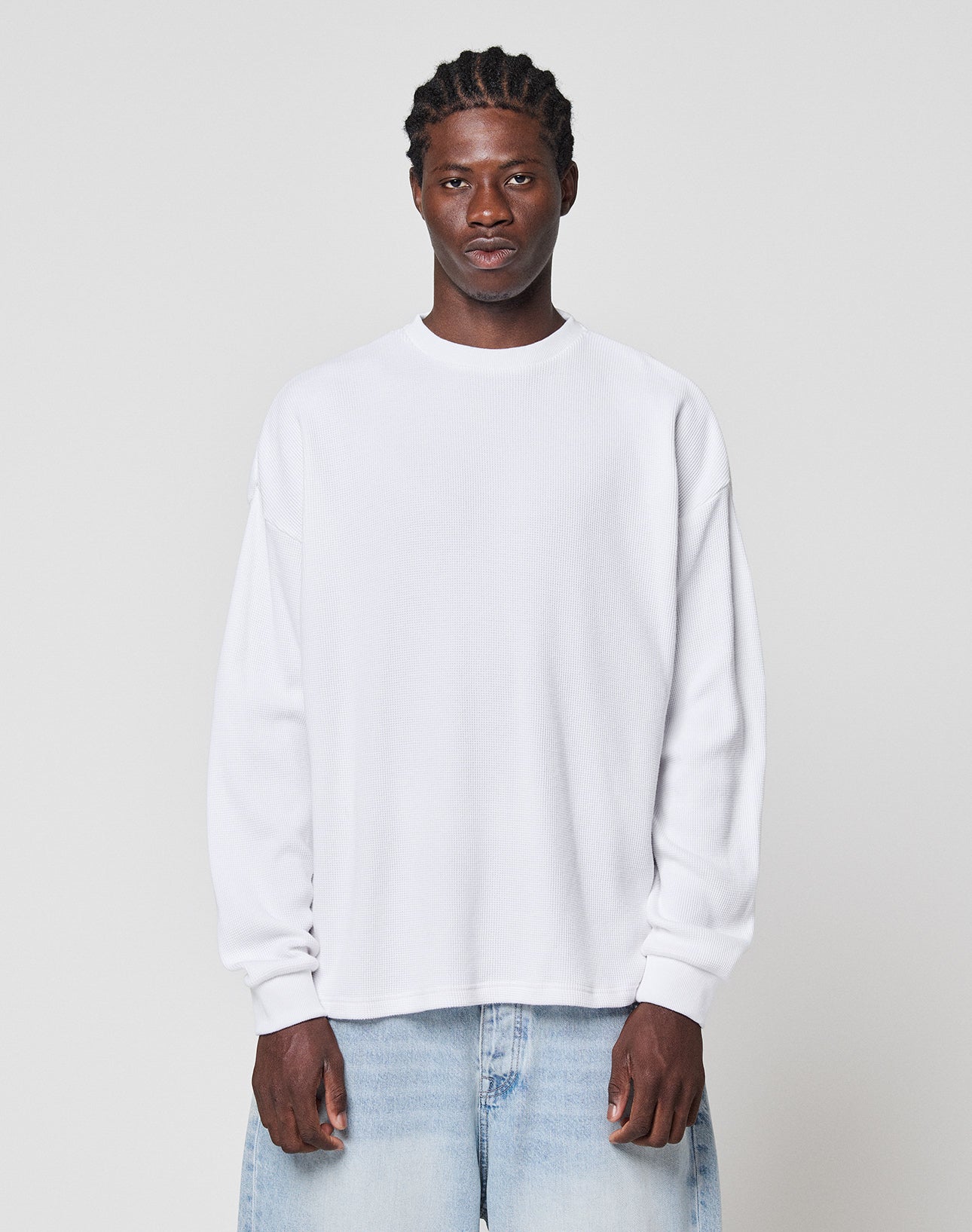 A man with braided hair wears the LIVE FAST DIE YOUNG Blank Waffle Longsleeve in white and light blue Oversize Fit jeans, standing against a plain light gray background, facing the camera with a neutral expression. | model