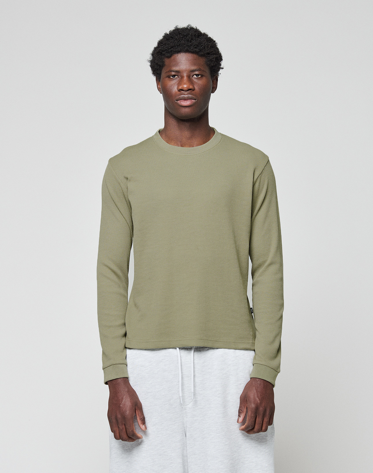A young man faces forward against a plain light gray background, wearing the LIVE FAST DIE YOUNG Basic Waffle Longsleeve in olive green and light gray sweatpants. | model