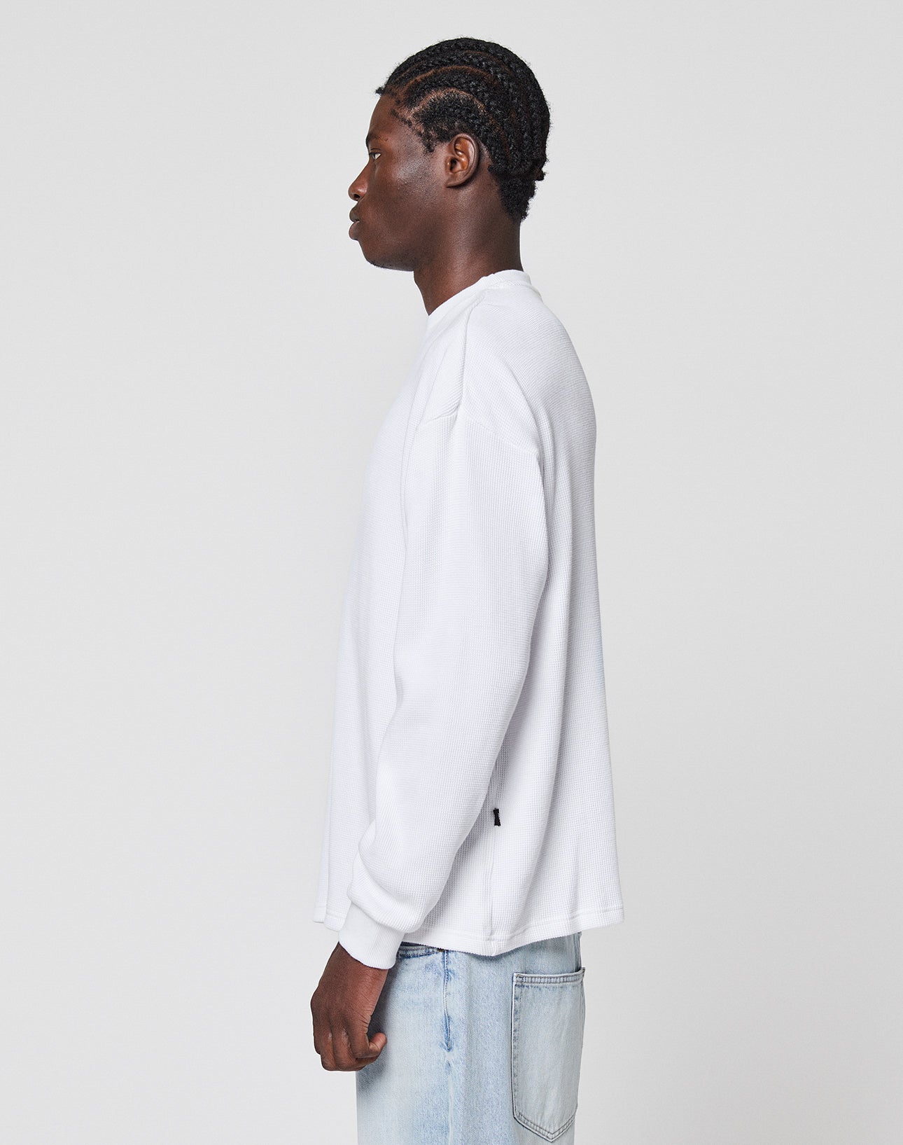 A person with braided hair wears the LIVE FAST DIE YOUNG Blank Waffle Longsleeve in oversize fit and light blue jeans, standing in profile facing left against a plain white background.