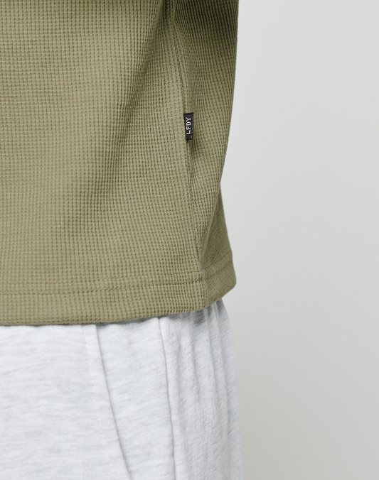 Close-up of a person wearing the olive-green LIVE FAST DIE YOUNG Basic Waffle Longsleeve in Oversize Fit with a small black flag label, paired with light grey pants against a plain, light background.