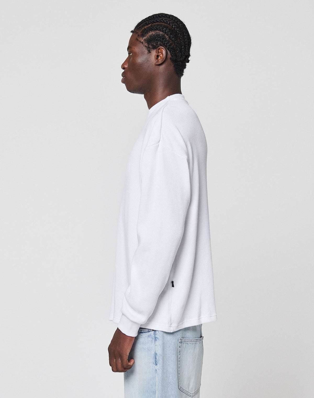 A person with braided hair wears the LIVE FAST DIE YOUNG Blank Waffle Longsleeve in oversize fit and light blue jeans, standing in profile facing left against a plain white background.