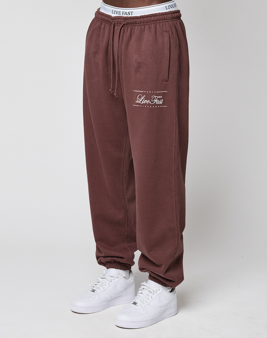 A person stands against a plain white background wearing LIVE FAST DIE YOUNG Wide Cuff Jogger in maroon with a white elastic waistband and white sneakers. | model