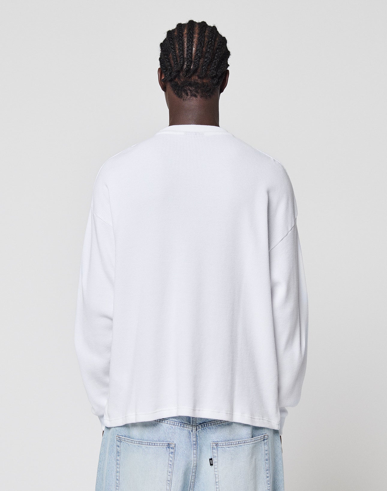 A person with braided hair wears the LIVE FAST DIE YOUNG Blank Waffle Longsleeve in oversize white (100% Baumwolle) and light blue jeans, standing with their back to the camera against a plain light gray background.
